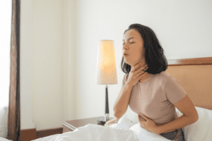Alt text: Mulher deitada na cama, com expressão de dor na garganta, representando o significado espiritual da laringite.